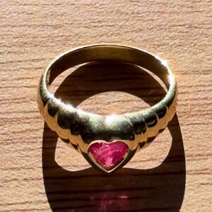 Pu&ograve; includere: Un anello in oro con una pietra preziosa rosa a forma di cuore incastonata al centro. La fascia presenta un design testurizzato e scanalato che si assottiglia verso il retro. Il metallo ha una finitura lucida che riflette la luce.