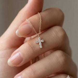 May include: A delicate gold necklace featuring a small cross pendant. The cross is adorned with sparkling, clear gemstones. The necklace is held up by a slender gold chain, creating a simple yet elegant piece of jewelry.