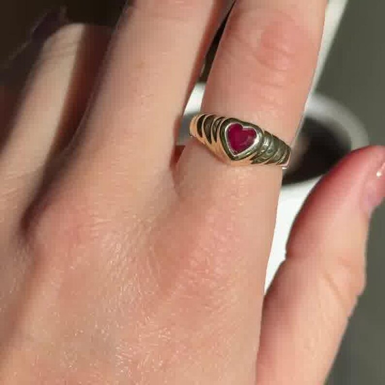 Pu&ograve; includere: Un anello in oro con una pietra preziosa rossa centrale a forma di cuore. La fascia presenta un design a coste testurizzato che si assottiglia verso il retro. Il metallo ha una finitura lucida e la pietra rossa &egrave; incastonata nel cuore.