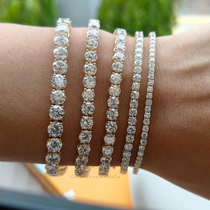 Könnte beinhalten: Fünf goldene Tennisarmbänder in unterschiedlichen Breiten, jedes mit einer durchgehenden Reihe runder, klarer, facettierter Steine. Die Armbänder sind nebeneinander angeordnet und zeigen ein abgestuftes Design vom breitesten zum schmalsten Band.