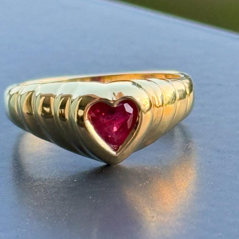 Pu&ograve; includere: Un anello in oro con una prominente pietra preziosa rossa a forma di cuore incastonata al centro. La fascia dell'anello presenta un design a coste testurizzato che avvolge il dito, creando un motivo continuo di linee in rilievo.