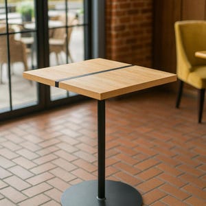 Könnte beinhalten: Ein quadratischer, hellfarbiger Holztisch mit schwarzem Metallsockel und einer zentralen Stütze. Die Tischplatte hat eine dunkle Akzentlinie. Der Tisch ist etwa 60 cm breit und eignet sich für ein Café oder Restaurant.