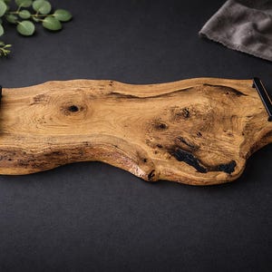 May include: A natural wood serving board with a live edge and black metal handles. The board is approximately 20 inches long and has a unique, organic shape. The wood grain is visible, showcasing the natural beauty of the material. Perfect for serving appetizers or charcuterie.