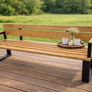 Op de afbeelding: Houten bank met een lichtbruine afwerking en zwarte metalen poten. Twee witte mokken en een kleine vaas met bloemen staan op een ronde houten dienblad. De bank staat buiten op een houten terras.