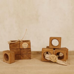 Puede incluir: Servilleteros de madera con acabado natural. Los anillos cuadrados tienen una abertura circular en el centro. Algunos están apilados, y otros están dispuestos con hilo y una etiqueta que dice "Handmade with love."