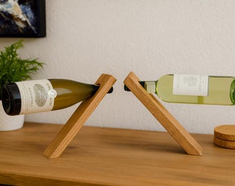 May include: Two wooden wine bottle holders, each supporting a bottle. One bottle is green with a white label, the other is a clear bottle with a white label. The holders are on a wooden surface.