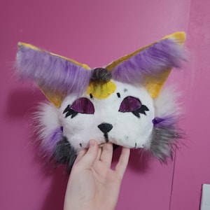 May include: A handmade cat mask in white, with purple, yellow, and grey details. The mask has large, pointed ears with purple fur, and purple mesh eyes. The nose and mouth are black. The mask is held by a hand.