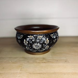May include: A decorative black ceramic bowl with a brown rim and base. The bowl features a white and gold floral pattern. The bowl is sitting on a wooden surface.