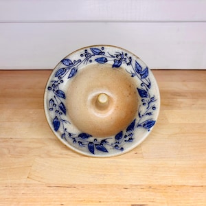 Op de afbeelding: Een ronde keramische ringhouder met een beige gespikkelde afwerking en een centrale pin. De brede, platte rand is voorzien van een handgeschilderd blauw botanisch patroon van fijne blaadjes en kleine besjes in een cirkelvormig rankmotief.