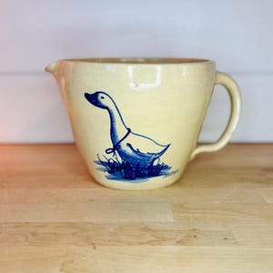 以下が含まれることがあります： クリーム色のセラミックピッチャー。青いガチョウのイラストが描かれています。ガチョウは青いリボンをしており、青い草の中に立っています。ピッチャーにはハンドルと注ぎ口が付いています。