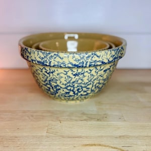 May include: A set of three nesting ceramic bowls. The bowls have a cream-coloured base with a blue spatter pattern. The bowls are stacked on a light wood surface.
