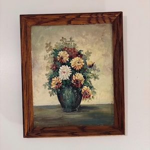 May include: Oil painting of a floral arrangement in a dark green vase, set against a light background. The flowers are a mix of white, yellow, and red, with green foliage. The painting is framed in a dark wood frame.