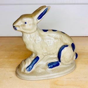 Może przedstawiać: Ceramiczna figurka królika w pozycji siedzącej, pokryta błyszczącym kremowym szkliwem z ręcznie malowanymi kobaltowymi akcentami na uszach, oczach, nosie, grzbiecie i łapach, umieszczona na zintegrowanej owalnej podstawie o gładkiej powierzchni.