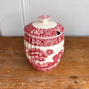 May include: A ceramic lidded jar featuring a red and white transferware pattern of floral borders and a landscape scene with trees. The jar has a ribbed body, a matching lid with a floral finial, and a small notch for a spoon on the rim.