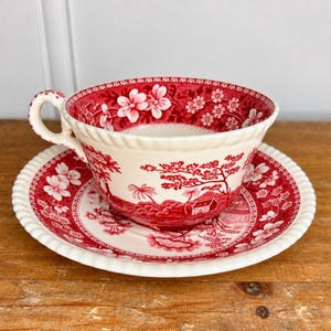 May include: A ceramic teacup and saucer set featuring a red and white transferware pattern. The design depicts detailed floral motifs and a landscape scene with trees and a small house. Both pieces have a scalloped, ribbed edge along the rim.