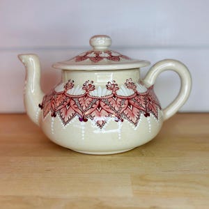 May include: Cream-colored ceramic teapot with a lid and handle. The teapot is decorated with a red and white floral pattern around the body and on the lid. The spout and handle are curved.