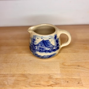 May include: A small, cream-colored ceramic creamer with a blue and white landscape design. The design depicts a barn or building with trees and birds. The creamer has a handle and a spout.