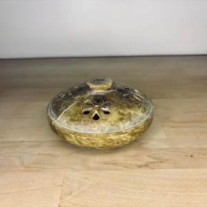 May include: A decorative, oval-shaped incense burner with a lid. The burner is a mottled gold and brown color, with a floral design on the lid and small holes for releasing the incense smoke. The burner sits on a wooden surface.