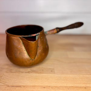 May include: A copper-colored, antique Turkish coffee pot with a wooden handle. The pot has a spout for pouring and is set on a light-colored wooden surface. The handle is dark brown and appears to be aged.