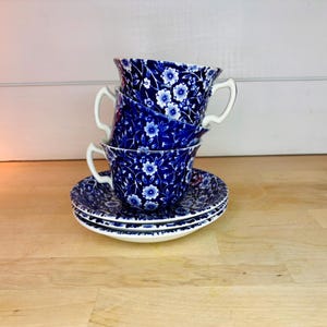 May include: A stack of two ceramic teacups and two matching saucers featuring a deep blue and white floral pattern. The teacups have white handles and a repeating botanical design of small blossoms and leaves across the entire exterior surface.