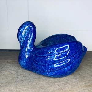 Puede incluir: Una maceta de cerámica con forma de cisne en un vibrante tono azul. La maceta presenta un diseño detallado con alas texturizadas y un cuello curvado. El interior es de un azul profundo, perfecto para pequeñas plantas o elementos decorativos.