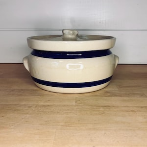 Puede incluir: Olla de cerámica color crema con tapa y dos asas. La olla presenta dos rayas azul marino alrededor del cuerpo. La tapa tiene un pequeño pomo para facilitar la extracción. La olla está sobre una superficie de madera.