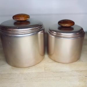Könnte beinhalten: Zwei passende Metallbehälter mit Deckeln. Die Behälter haben eine gebürstete Kupferoberfläche und sind zylindrisch geformt. Jeder Deckel ist mit einem kleinen, runden Holzknopf versehen. Die Behälter sind unterschiedlich groß.