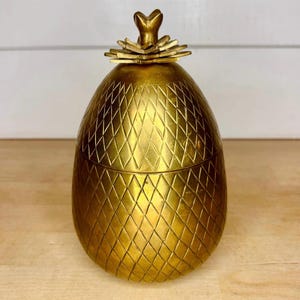 Puede incluir: Un recipiente de metal dorado con forma de piña y un patrón de diamantes texturizado en toda su superficie. El objeto cuenta con una tapa extraíble rematada con un remate de hojas superpuestas y estilizadas, creando una silueta redondeada.
