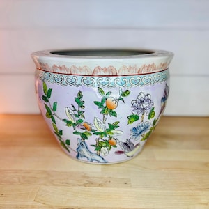 May include: A round, ceramic planter with a light purple background and floral and bird designs. The planter has a wide rim with a red and white pattern. The floral design includes orange fruits, green leaves, and blue and white flowers.
