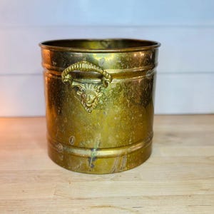 Peut inclure: Seau en laiton vintage avec une poignée décorative ornée d'une tête de lion. Le récipient cylindrique a un aspect vieilli et terni, suggérant qu'il s'agit d'une antiquité. Le seau est posé sur une surface en bois.