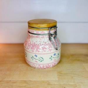 May include: A ceramic storage jar with a light pink speckled pattern and a horizontal band featuring small green and blue floral motifs. The jar includes a wooden lid secured by a silver-toned metal wire bail closure mechanism.