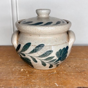 Puede incluir: Una vasija de cerámica beige moteada con tapa a juego y dos asas laterales. El exterior presenta motivos de hojas pintados en verde oscuro y una marca rectangular estampada que dice SALMON FALLS POTTERY 1999 O.R.S. en el centro del cuerpo.