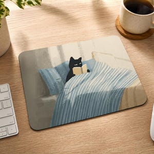 May include: A rectangular mousepad featuring a black cat reading a book in bed. The cat is under a blue and white striped blanket, with a blue pillow. The background is a light beige color. A white keyboard and mouse are visible.