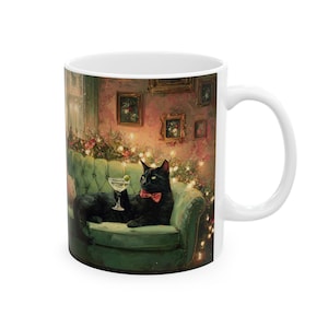 Può includere: Una tazza in ceramica bianca con un'illustrazione stampata di un gatto nero con papillon rosso, sdraiato su un divano verde imbottito mentre tiene in mano un bicchiere da martini in una stanza con luci calde.