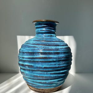 Puede incluir: Jarrón de cerámica turquesa hecho a mano con textura acanalada horizontal. El jarrón tiene un borde y una base marrones, añadiendo un elemento terroso contrastante. Ideal para flores o como objeto de arte. Altura aproximada: 25 cm.