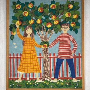 Può includere: Opera d'arte tessile incorniciata raffigurante un melo stilizzato con mele rosse e gialle. Una ragazza con un vestito giallo e un ragazzo con una camicia a righe rosse sono vicino all'albero. Una staccionata rossa e uno sfondo blu completano la scena.