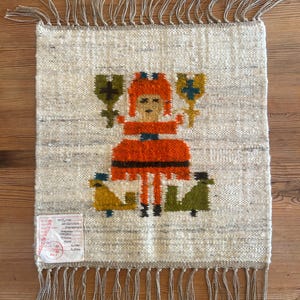 Può includere: Arazzo tessuto con un design di arte popolare. La figura centrale è una persona in un vestito arancione, che tiene due oggetti floreali stilizzati. Lo sfondo è beige chiaro con frange in alto e in basso. Un'etichetta è attaccata in basso a sinistra.