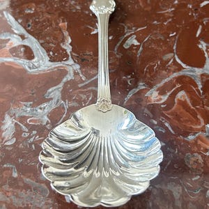 Puede incluir: Una cuchara de servir plateada con un cuenco en forma de concha y un mango decorativo. El cuenco de la cuchara tiene un borde festoneado y un diseño acanalado. El mango tiene un diseño floral adornado en la parte superior. La cuchara es probablemente de plata de ley.