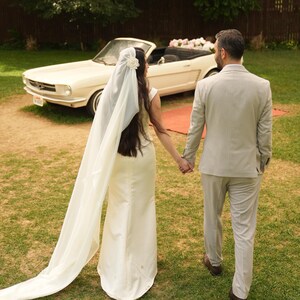 May include: A bride and groom walking away from a white vintage car with a floral arrangement on the back seat. The bride is wearing a white wedding dress and a long white veil. The groom is wearing a light grey suit.