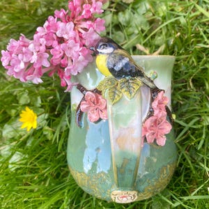 Puede incluir: Una taza de cerámica verde con un pájaro y flores esculpidas en el asa. La taza está sentada sobre un lecho de hierba verde con flores rosas.