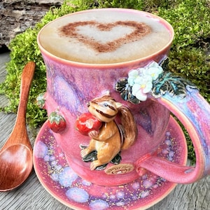 Puede incluir: Un juego de taza y platillo de café de cerámica rosa con un diseño caprichoso de ardilla. La taza contiene un capuchino con un diseño en forma de corazón. Una cuchara de madera descansa junto a la taza.