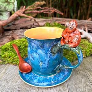 Pode incluir: Uma chávena de café de cerâmica azul com uma pega em forma de orangotango. O interior da chávena é amarelo e assenta num pires a condizer. Uma pequena colher de madeira repousa sobre o pires. A chávena tem cerca de 10 cm de altura.