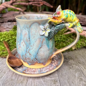 Könnte beinhalten: Eine Keramiktasse mit blau-brauner Glasur, mit einem gelb-grünen Chamäleon auf dem Griff. Eine weiße Blume und grüne Blätter zieren den Griff. Die Tasse steht auf einer passenden Untertasse mit einem Holzlöffel.