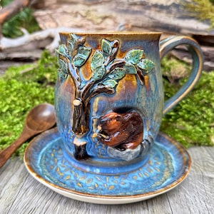 Puede incluir: Un juego de taza y platillo de cerámica hecho a mano. La taza presenta un relieve detallado de un árbol y un castor. El juego está esmaltado en tonos azules, marrones y verdes. Una cuchara de madera descansa cerca.