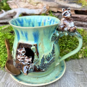 Op de afbeelding: Een turquoise keramische mok met blauwe accenten, met een wasbeerontwerp. De handgreep heeft een wasbeerfiguur en de mok heeft drie wasberen in een boomholte. Inclusief bijpassende schotel en houten lepel. Uniek ontwerp.