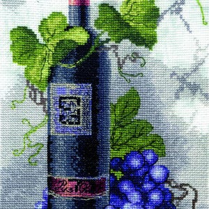 May include: A cross-stitch embroidery depicting a dark wine bottle with a pink foil top, a purple label, and a red label strip. A cluster of blue grapes, green vine leaves, and a small red ladybug are positioned around the bottle on a light surface.
