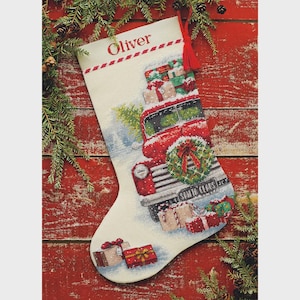 May include: A cream-colored Christmas stocking featuring a cross-stitch design of a red vintage truck carrying wrapped gifts and a wreath. The name Oliver is embroidered in red at the top. A red tassel hangs from the top corner of the stocking.