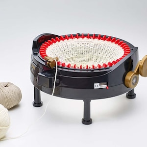 Pode incluir: Uma máquina de tricotar circular preta com uma base de agulhas vermelha e uma manivela dourada, apresentando um tubo de malha branco que emerge do centro. Três novelos de lã em tons de creme, bege e castanho estão perto das pernas.