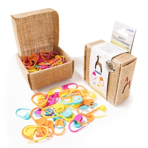 Pode incluir: Um conjunto de marcadores de pontos de segurança da marca Pony numa caixa de juta. Os marcadores são de plástico, de forma oval e vêm em laranja, amarelo, rosa e azul. A embalagem inclui uma etiqueta com o texto Safety Stitch Markers x 40 e SA 8000 Certified.