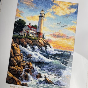 Puede incluir: Un kit de punto de cruz contado Dimensions Gold Collection titulado Rocky Point. El empaque muestra un faro en un acantilado rocoso sobre un océano agitado al atardecer, con aves volando en un cielo amarillo y azul sobre el agua.